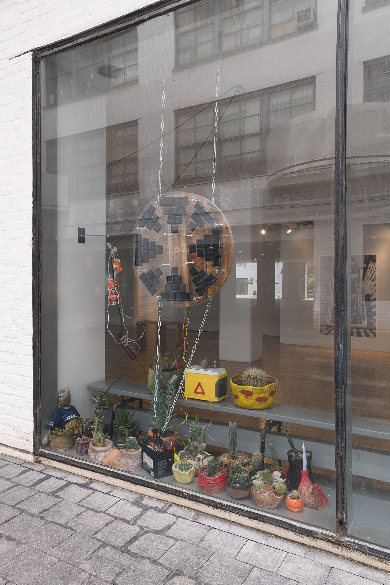 [Exterior view of floor-to-ceiling gallery windows from courtyard. Through the window you can see a sculptural installation. On the floor there are around twenty colorful, handmade ceramic planters with various types of cacti. Hanging from two parallel silver chains above them is a flat, circular piece of wood with triangular configurations of small solar panels on the face of it.]