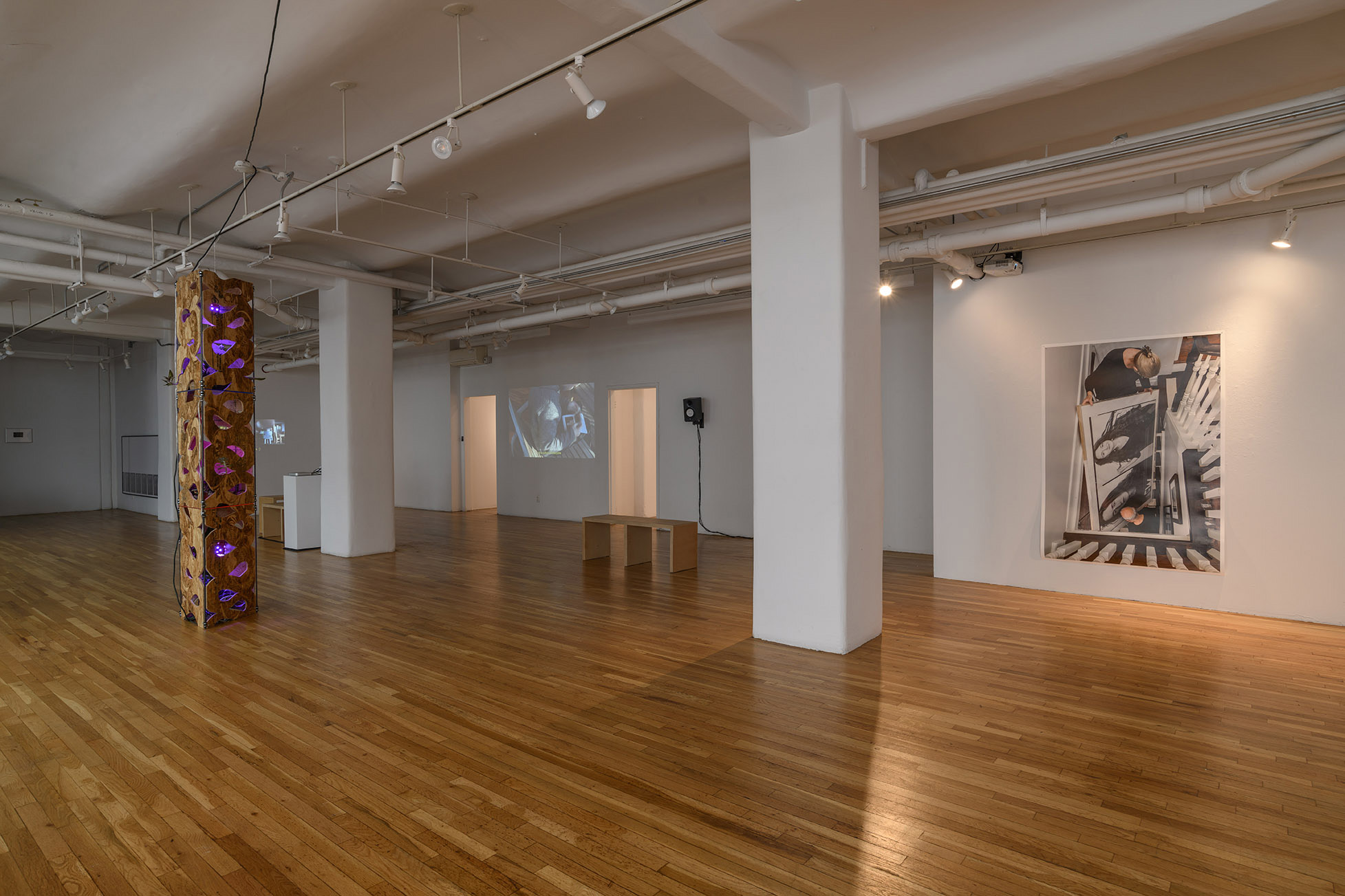 [A large open gallery with wooden flooring supported by two white concrete columns. Stationed near the two columns, almost midway between them, is a tall column-like wooden sculpture that nearly reaches the ceiling. The gallery’s lighting track is primarily turned off except for lights illuminating a large unframed color photograph hanging on a wall opposite the column-like wooden sculpture. Receding in space on the same side of the gallery as the photograph is a single speaker monitor mounted high on a white wall, two open doorways illuminated by warm light, and color video projections either side of the illuminated doorways.]