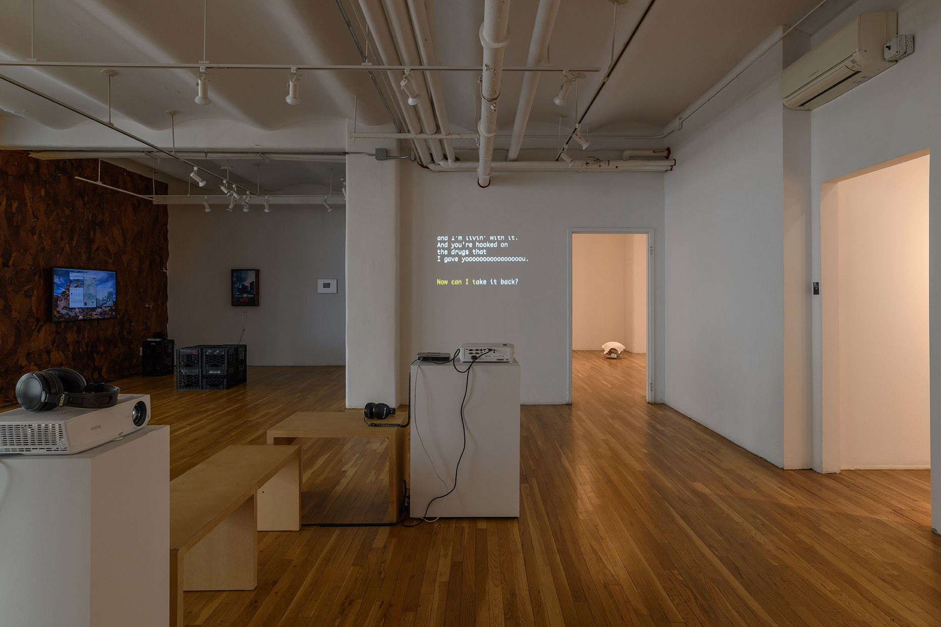[Another view of the large open gallery facing the rear of the gallery space. The center of this view shows two white plinths, each supporting a projector, cabling, and headphones. Between the plinths are two benches stationed perpendicular to one another. Farthest from the plinths and benches is a large, dimly lit alcove featuring sculptures on the floor and wall, a drawing, and a lit TV monitor. To the right of this alcove is a video projected onto a short wall connected to one of the illuminated doorways. Turning the corner is another white wall abutting another illuminated open doorway.]