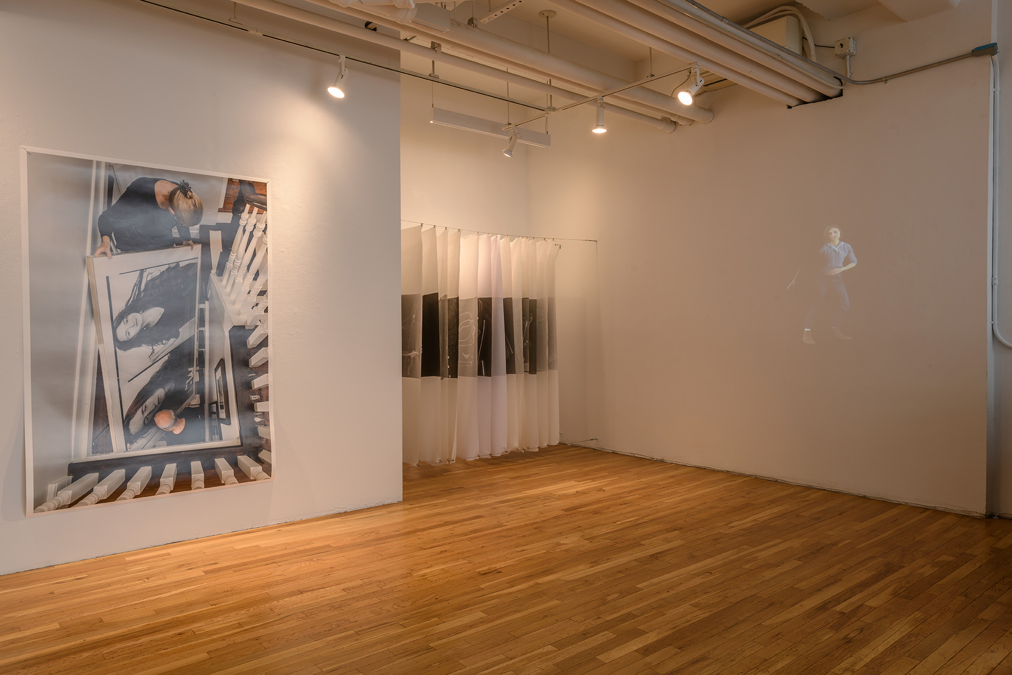[Another view of the large open gallery facing more directly the large wall-hung unframed color photograph. To the right of the photograph is a sculpture made of sheets of diaphanous fabrics of various opacities. Turning the corner from the fabric sculpture, a faintly-seen moving image is projected onto a white wall.]