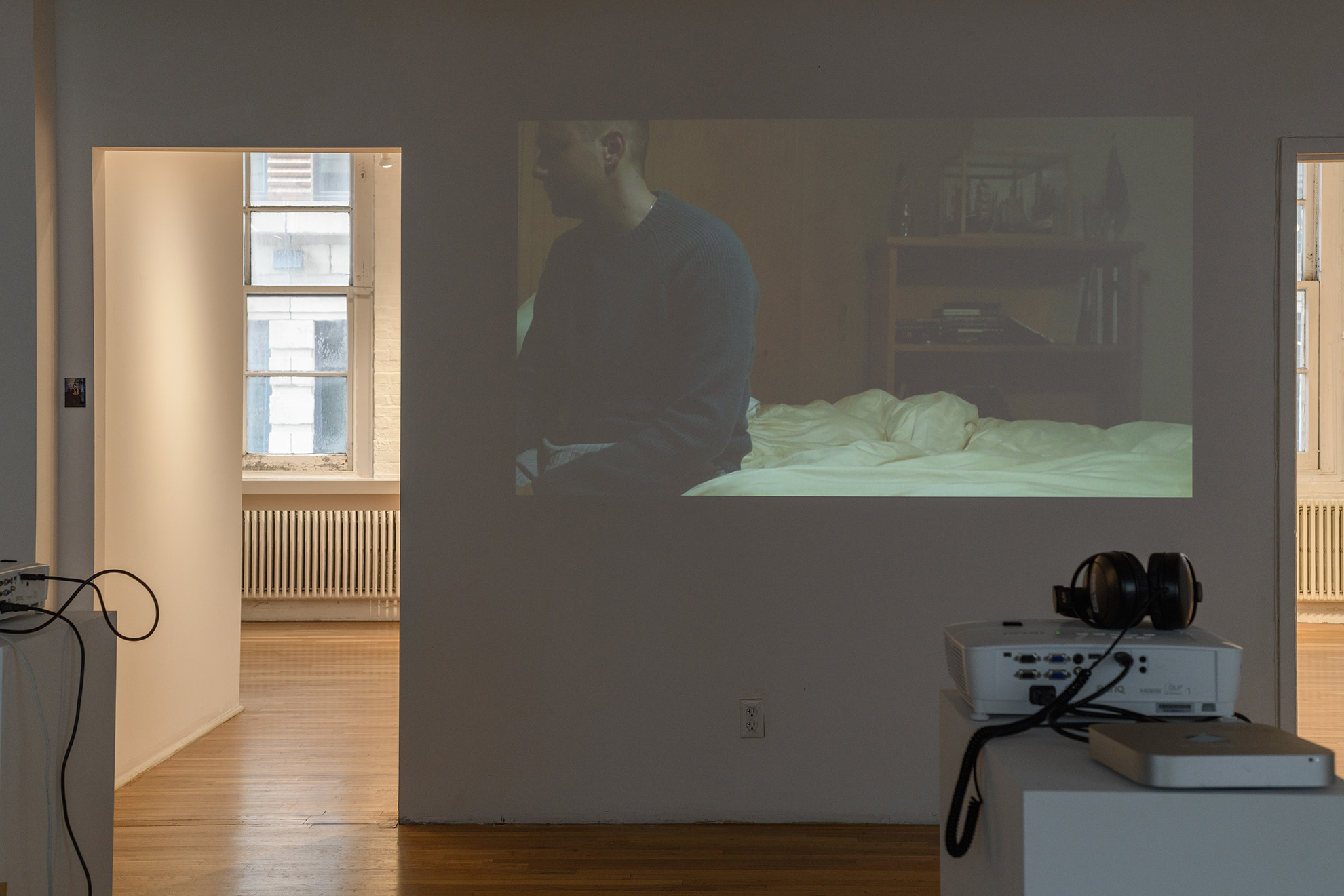 [Another view of the same projection at a different point in the video showing a person, who appears to be white, wearing a blue sweater and sitting at the edge of an unmade bed in a dim lit room, gazing outside of the frame. On either side of the projected image are doorways leading to additional galleries.]