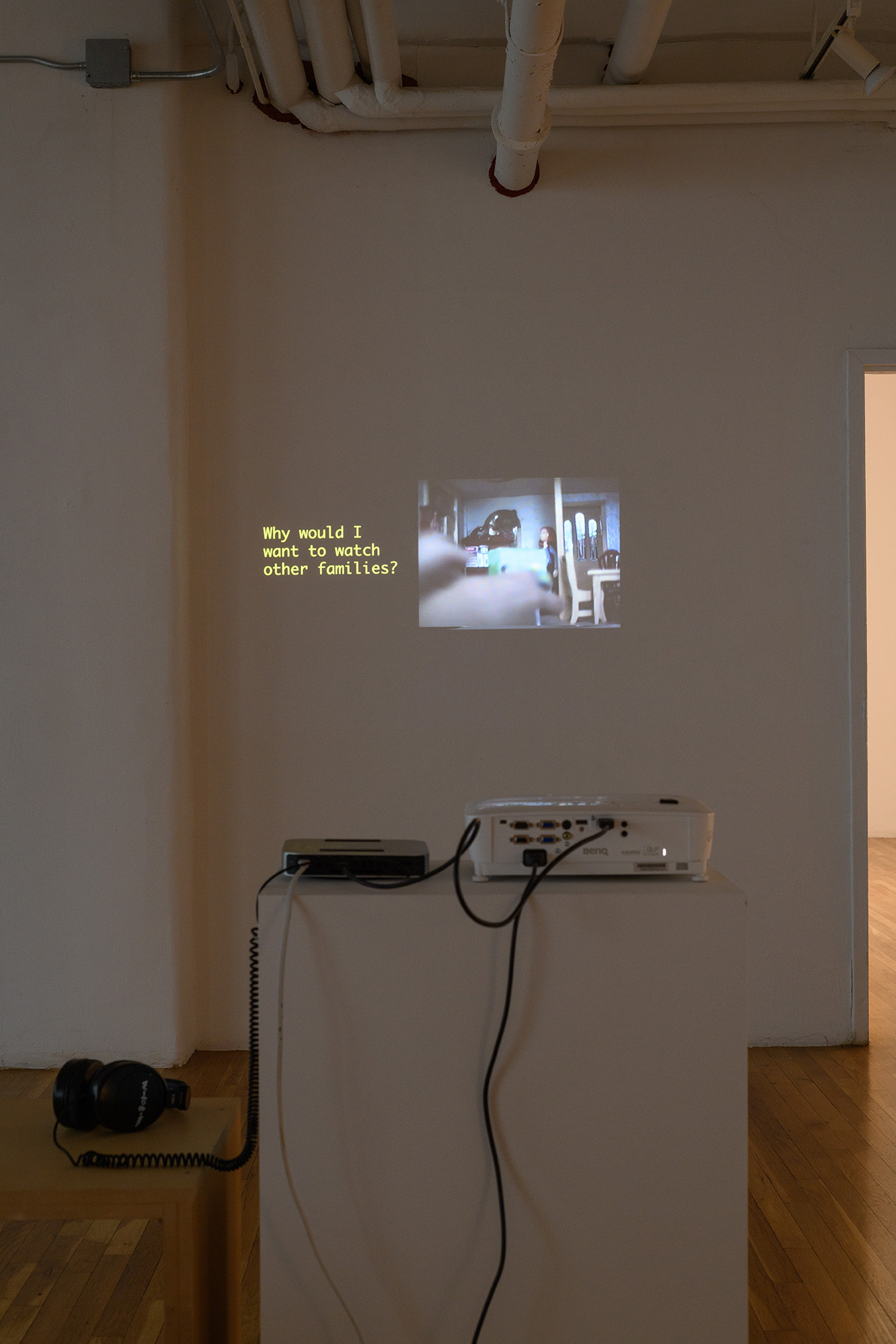 [A video projection. Both the projected image and the projector machine, media player, and cabling, all sit on a white plinth. The projected image shows a small image of the interior of a doll house with blurry hands in the foreground. To the left of the image, yellow text reads, quote: “Why would I want to watch other families?”]