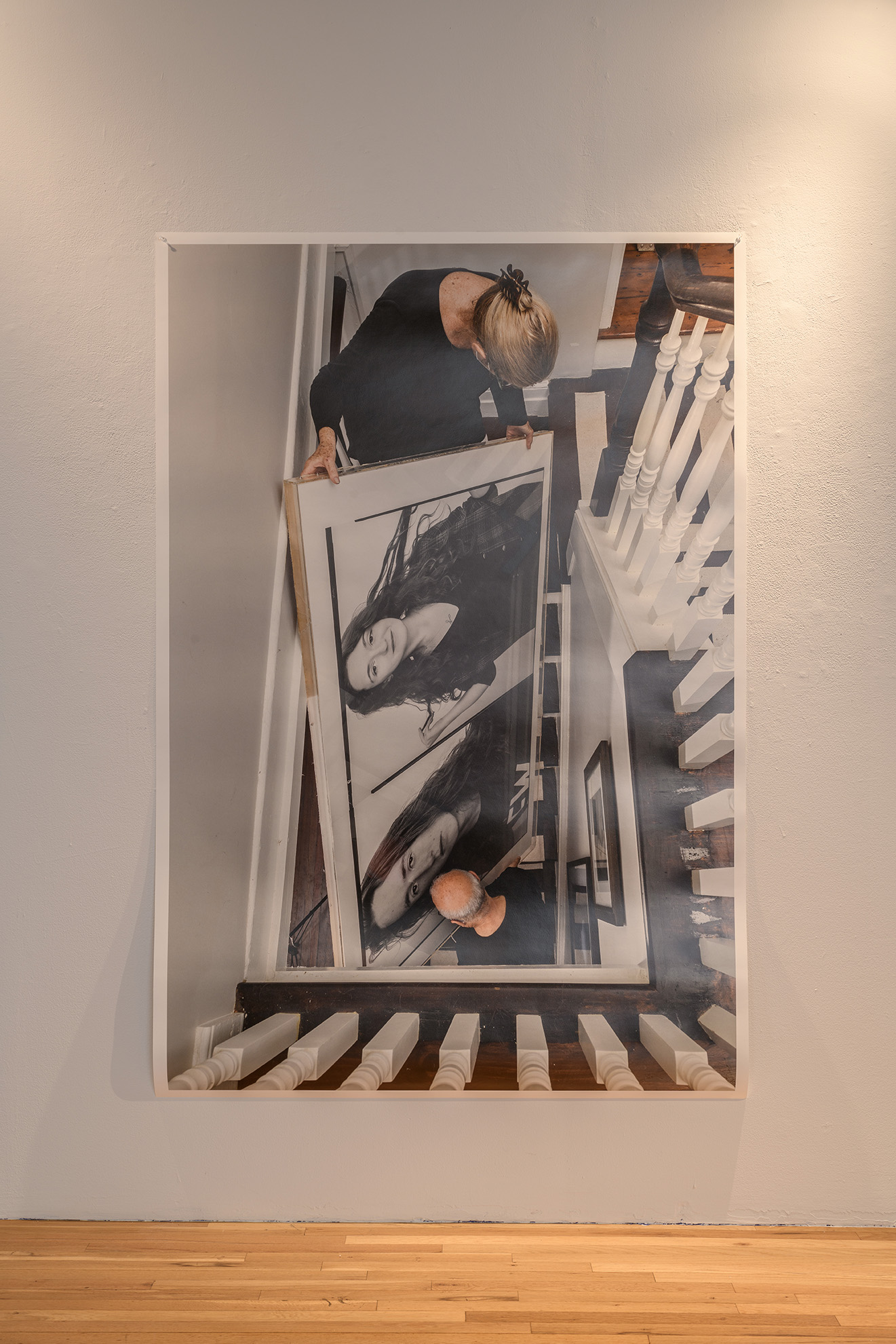 [An unframed color photograph depicting two people carrying a large artwork down a steep, narrow staircase. The artwork being carried is a large framed photograph of two studio portraits. All of the figures depicted, both the people carrying the photograph and the portraits of the people photographed appear to be white and the two people carrying the framed photograph appear to be much older than the two people in the portrait.]