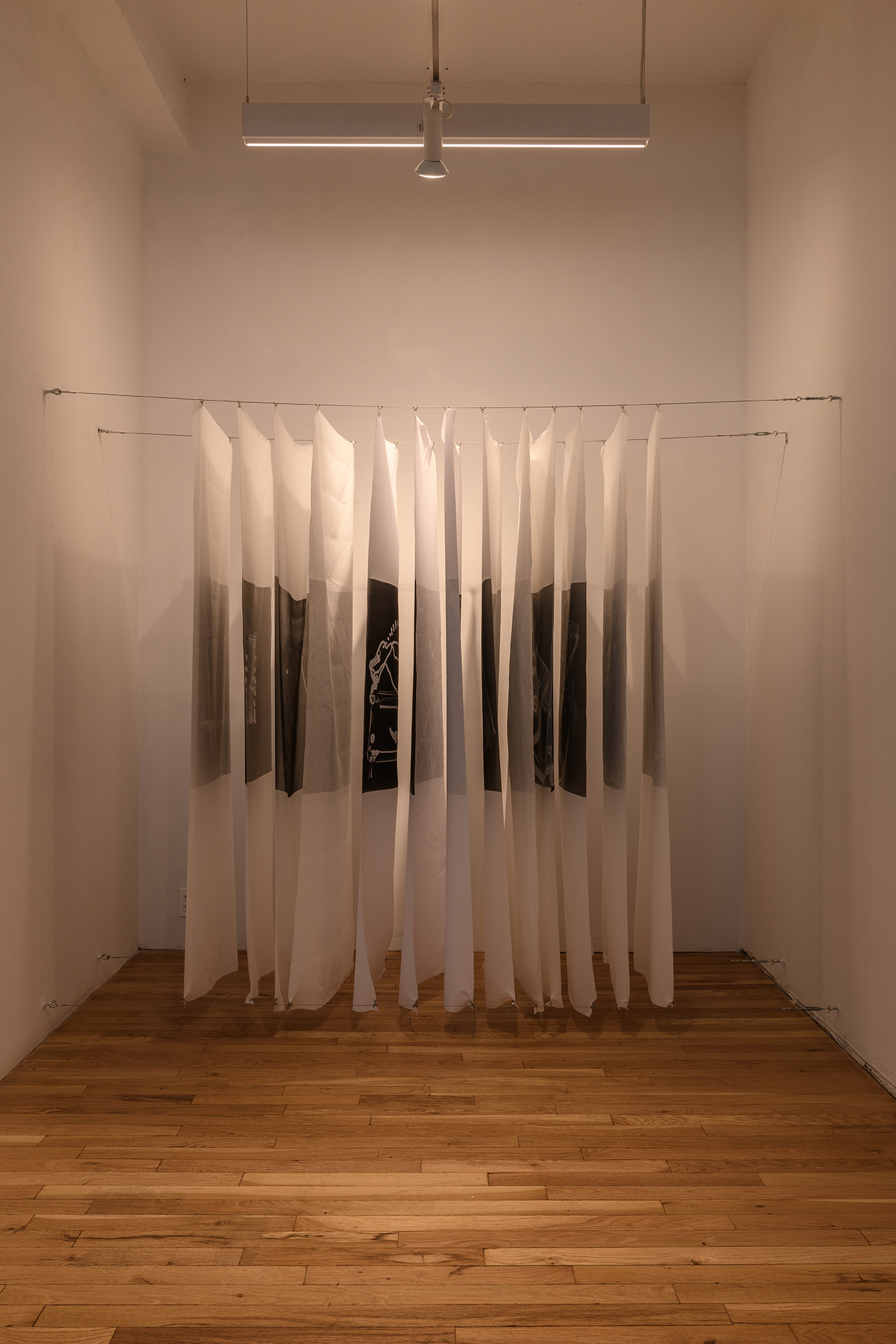 [In an alcove of the gallery, orderly sheets of silk are held in an evenly spaced stack like slices of bread. An X-ray image of clothing or fashion accessories is printed in a dark rectangle at the center of each sheet. One of the sheets folds open and reveals an x-ray of a handbag. The metal fixtures on the bag stand out as bright white zippers and chains as the fibrous material of the bag blends with the black background of the image.]