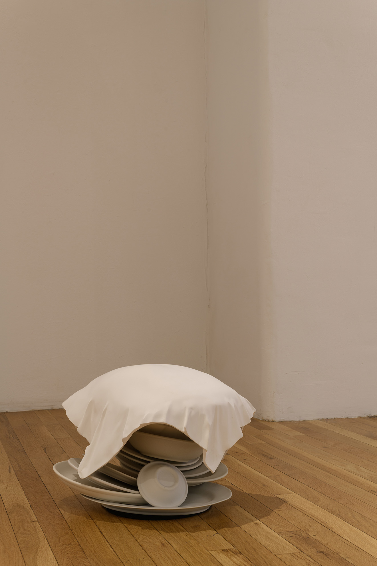 [A cream-colored sculpture sitting directly on the floor. The sculpture appears to be a stack of dishes with a cloth draped over them. Plates, bowls and platters. On closer inspection the cloth has the shape of a person's back, bending over as though picking something up off the floor. On even closer inspection, the back-shaped-cloth turns out to be a solid material closer in makeup to the dishes it covers than to textile or flesh.]