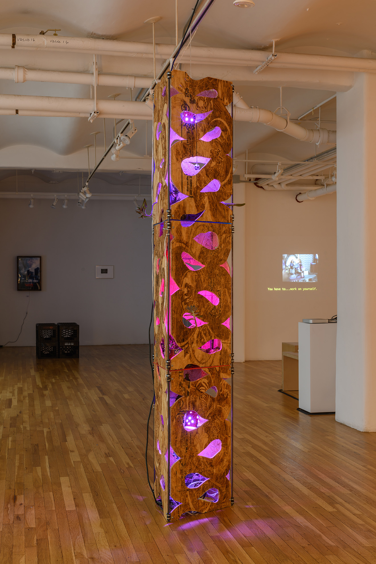 [A tall, totem-like sculpture is illuminated with pink and purple lights from within. The work is made of CNC’d and laser etched plywood planks. Cuts in the wood allow areas of light to leak out of the sculpture.]